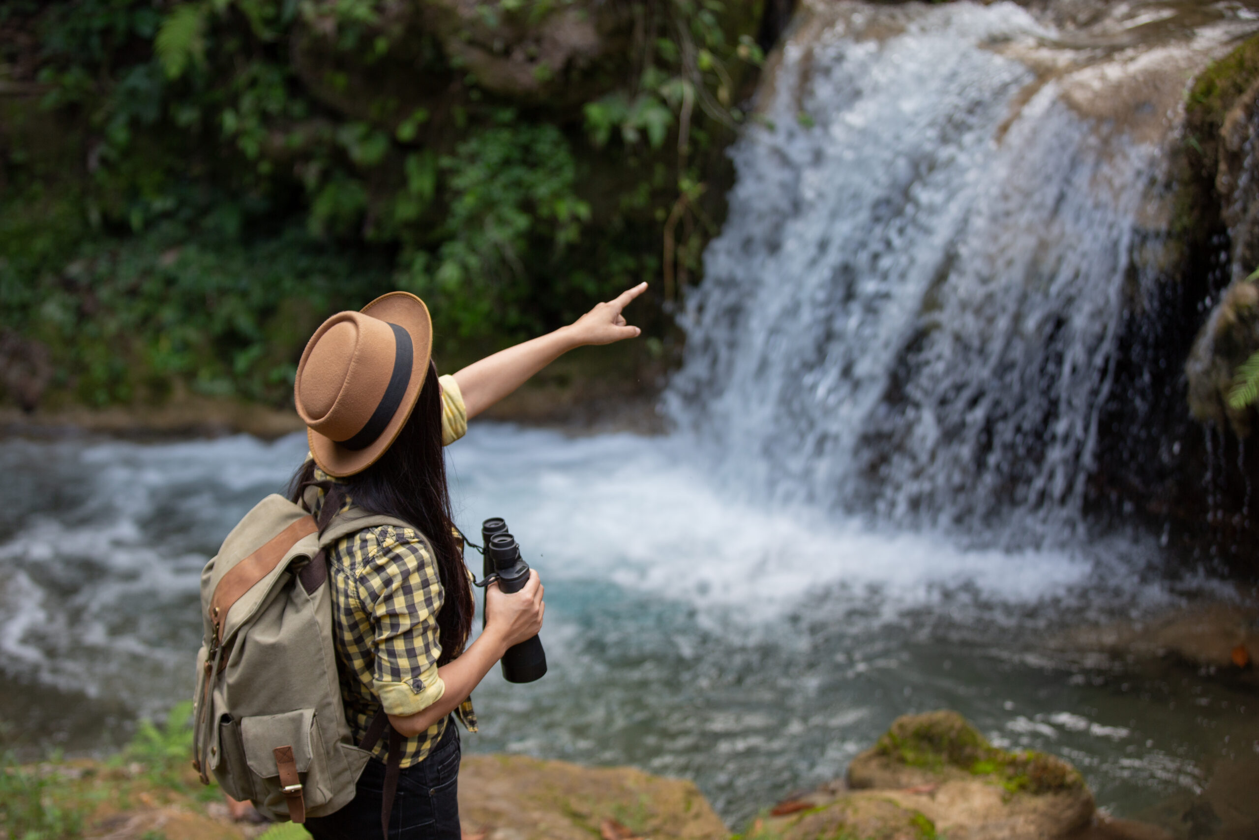 Mujer con sombrero y mochila observando una cascada en un entorno natural, simbolizando el turismo sostenible y la conexión con la naturaleza.
